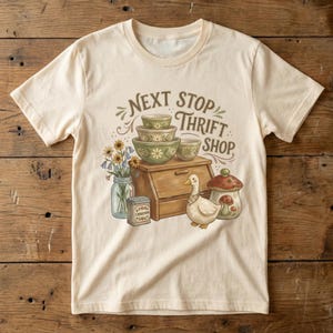 May include: Cream-colored t-shirt with a graphic featuring a wooden cabinet, bowls, a duck, and a mushroom-shaped jar. The text "Next Stop Thrift Shop" is arched above the cabinet. A jar of flowers and a cinnamon tin are also included.