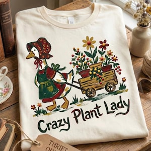 Puede incluir: Camiseta color crema con una ilustración caprichosa de un ganso tirando de un carro de madera lleno de macetas con flores. El ganso lleva un gorro y un delantal. El texto en la camiseta dice "Crazy Plant Lady".