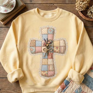 Puede incluir: Una sudadera amarilla clara con un dise&ntilde;o de cruz de retazos. La cruz est&aacute; adornada con patrones florales y de cuadros, un ribete de encaje y un ramo de flores atado con un lazo. La sudadera tiene mangas largas y cuello redondo.