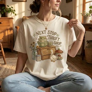 May include: Cream-colored t-shirt with a graphic that reads "Next Stop Thrift Shop." The graphic features stacked bowls, a wooden breadbox, a duck, and a mushroom. The shirt is worn by a person sitting on a rug.