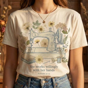 Könnte beinhalten: Cremefarbenes T-Shirt mit einer detaillierten Illustration einer Vintage-Nähmaschine, umgeben von floralen Elementen. Der Text "She works willingly with her hands" und "Proverbs 31:13" ist darunter gedruckt.