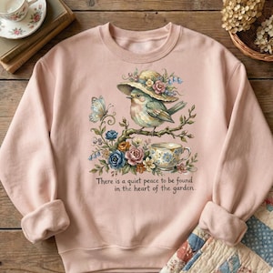 Pu&ograve; includere: Una felpa rosa chiaro con un'illustrazione ad acquerello di un uccello con un cappello, appollaiato su un ramo con fiori e una tazza da t&egrave;. &Egrave; presente anche una farfalla. Il testo recita: "C'&egrave; una quieta pace da trovare nel cuore del giardino."