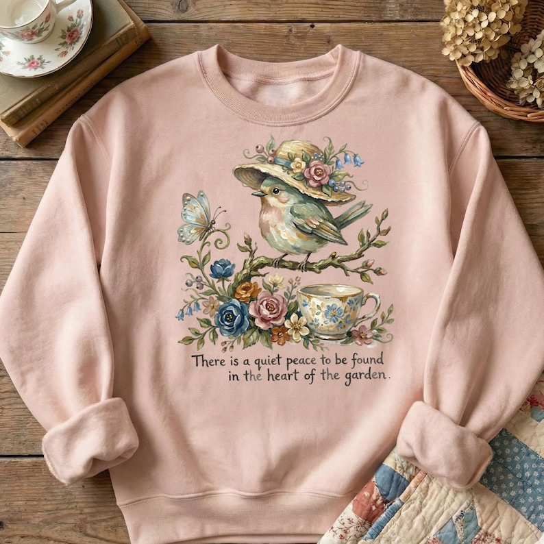Pu&ograve; includere: Una felpa rosa chiaro con un'illustrazione ad acquerello di un uccello con un cappello, appollaiato su un ramo con fiori e una tazza da t&egrave;. &Egrave; presente anche una farfalla. Il testo recita: "C'&egrave; una quieta pace da trovare nel cuore del giardino."