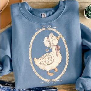 May include: A light blue sweatshirt featuring a whimsical illustration of a goose wearing a bonnet and a pink bow. The goose is inside an oval frame with a lace-like border. The goose has floral patterns on its body.
