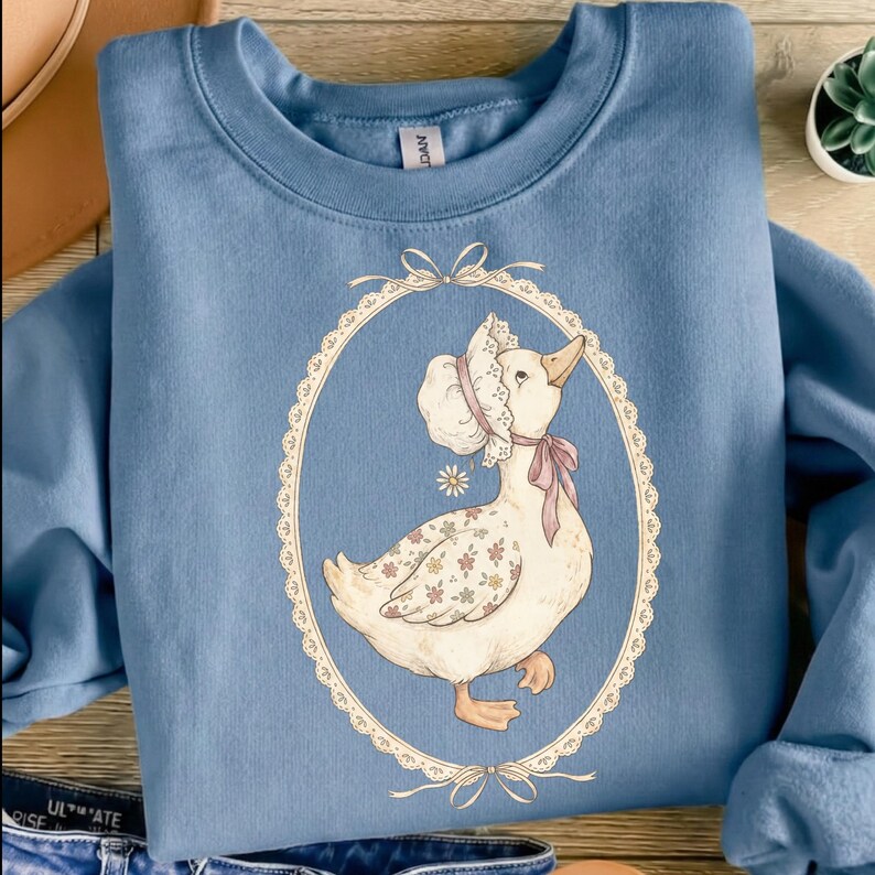 May include: A light blue sweatshirt featuring a whimsical illustration of a goose wearing a bonnet and a pink bow. The goose is inside an oval frame with a lace-like border. The goose has floral patterns on its body.