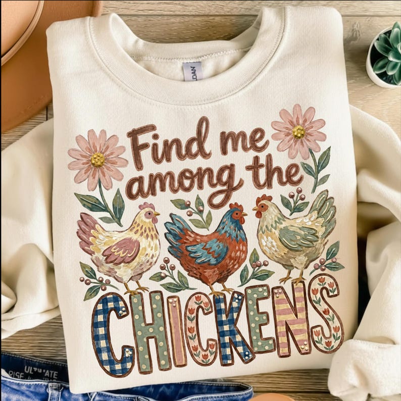 May include: A cream-colored crewneck sweatshirt featuring a graphic print of three chickens surrounded by pink flowers and green leaves. The text reads Find me among the CHICKENS, with the word CHICKENS in large, patterned, multi-colored letters.