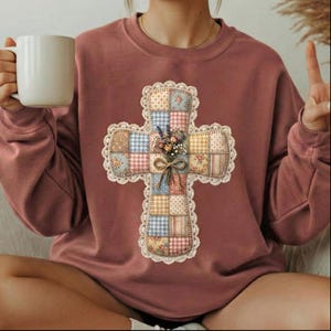 Puede incluir: Una sudadera de color malva con un dise&ntilde;o de cruz de retazos. La cruz est&aacute; adornada con patrones florales y de cuadros, un ribete de encaje y un peque&ntilde;o ramo. La sudadera de manga larga tiene cuello redondo.