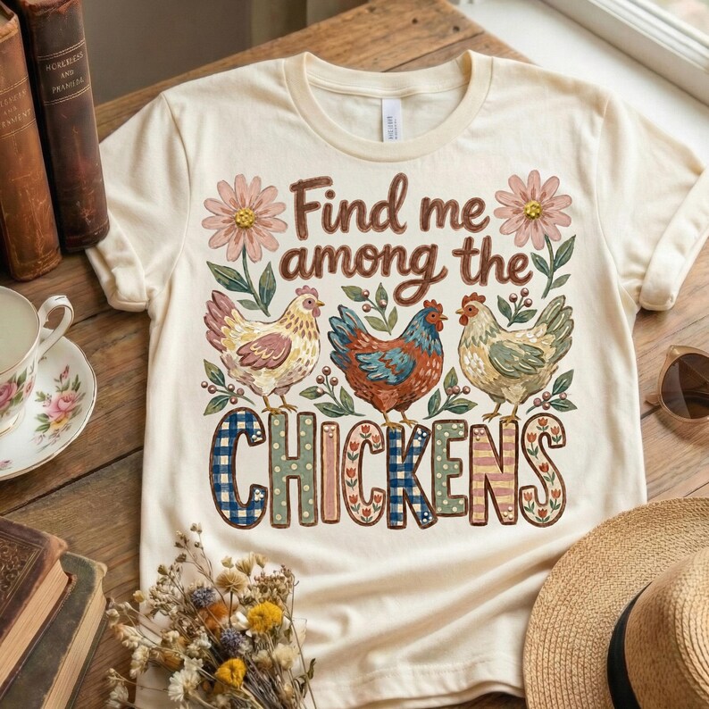May include: Cream-colored t-shirt featuring the text "Find me among the Chickens" with floral and chicken illustrations. The design includes three chickens in different colors and patterns, with the word "CHICKENS" in a mix of patterned fonts.