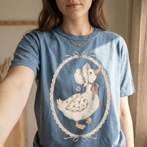 May include: A blue t-shirt featuring a vintage-style illustration of a goose wearing a bonnet and a pink bow. The goose is inside an oval frame with a lace-like border. The goose has floral patterned wings.