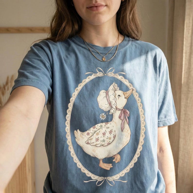 May include: A blue t-shirt featuring a vintage-style illustration of a goose wearing a bonnet and a pink bow. The goose is inside an oval frame with a lace-like border. The goose has floral patterned wings.