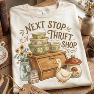 May include: Cream-colored t-shirt with the text "NEXT STOP THRIFT SHOP" and illustrations of bowls, a duck, a mushroom jar, and flowers. The design has a vintage, farmhouse aesthetic.