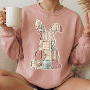 Puede incluir: Una sudadera rosa claro con un dise&ntilde;o de conejo de retazos. El conejo est&aacute; compuesto por varios patrones de tela, incluyendo estampados florales, a rayas y a cuadros. La sudadera tiene mangas largas y cuello redondo.
