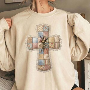 Puede incluir: Sudadera de color crema con un dise&ntilde;o de cruz de retazos. La cruz est&aacute; adornada con patrones florales y de cuadros, un ribete de encaje y un peque&ntilde;o ramo de flores en el centro. La sudadera tiene mangas largas y cuello redondo.