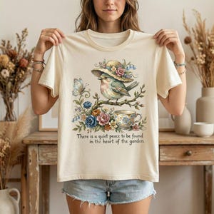 Pu&ograve; includere: T-shirt color crema con un'illustrazione ad acquerello di un uccello con un cappello, appollaiato su un ramo con fiori e una tazza da t&egrave;. Il testo "There is a quiet peace to be found in the heart of the garden" &egrave; stampato sotto.