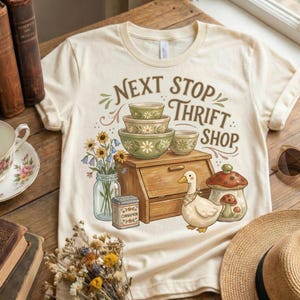 May include: Cream-colored t-shirt with the text "Next Stop Thrift Shop" and a vintage-style graphic. The graphic includes stacked green bowls, a wooden breadbox, a duck, and a mushroom. A jar of flowers and a cinnamon tin are also featured.