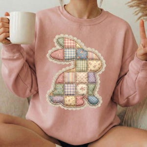 Pode incluir: Um moletom rosa claro com um design de coelho de retalhos. O coelho é feito de vários tecidos estampados, incluindo florais, xadrez e bolinhas, delineados com uma borda de renda. O moletom tem mangas compridas.
