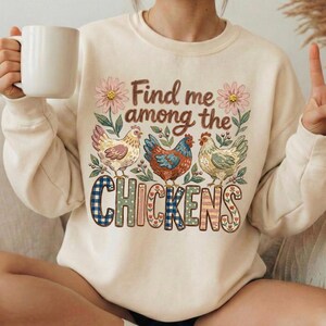 May include: Cream-colored sweatshirt with a floral and chicken graphic. The design features three chickens, pink flowers, and the text "Find me among the Chickens" in a decorative font. The word "CHICKENS" is in a plaid and floral pattern.