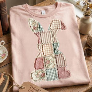Puede incluir: Una camiseta rosa claro con un dise&ntilde;o de conejo de patchwork. El conejo est&aacute; hecho de varios patrones de tela, incluyendo estampados florales, a rayas y a cuadros en tonos de rosa, verde y crema. La camiseta est&aacute; doblada sobre una superficie de madera.