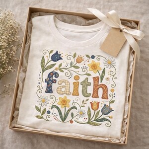 May include: A cream-coloured t-shirt featuring the word "faith" in a floral design, with daisies, daffodils, and tulips. The shirt is presented in a gift box, tied with a ribbon and a tag.