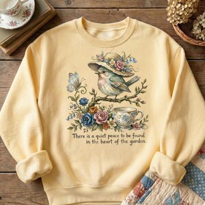 Pu&ograve; includere: Felpa giallo chiaro con un'illustrazione dettagliata di un uccello con un cappello, appollaiato su un ramo fiorito. Sono raffigurati anche una tazza da t&egrave; e una farfalla. Il testo "There is a quiet peace to be found in the heart of the garden" &egrave; sotto.