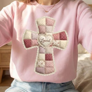 May include: A light pink sweatshirt featuring a cross design. The cross is made of patchwork squares in shades of pink, cream, and burgundy, with a heart in the center that says "Loved". The cross is trimmed with lace.