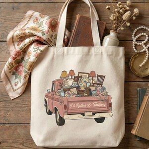 May include: Cream-colored tote bag featuring a pink vintage truck illustration filled with furniture and decor. The truck bed is overflowing with lamps, chairs, dishes, and blankets. The text "I'd Rather Be Thrifting" is printed on the truck. A floral scarf and books are visible.