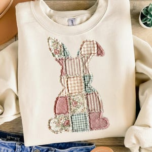 Könnte beinhalten: Cremefarbenes Sweatshirt mit einem Patchwork-Hasen-Design. Der Hase besteht aus verschiedenen Stoffmustern, darunter Blumen-, Streifen- und Vichy-Muster, in Rosa-, Grün- und Cremetönen. Das Sweatshirt hat einen Rundhalsausschnitt und lange Ärmel.