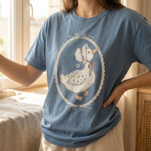 May include: A light blue t-shirt featuring a whimsical illustration of a goose wearing a bonnet and a pink bow, set within an oval frame. The goose has floral patterned wings. The frame is decorated with lace and bows.