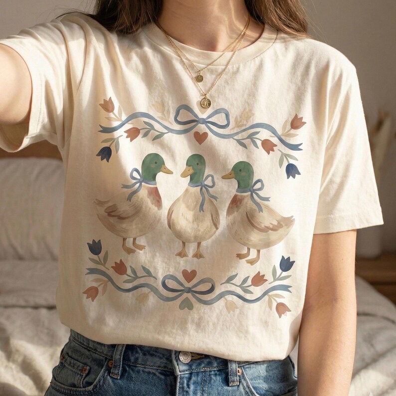 May include: Cream-coloured t-shirt featuring a watercolour design of three ducks with green heads and blue bows. The ducks are surrounded by a floral and ribbon border in blue, red, and green. The shirt is made of a soft material.