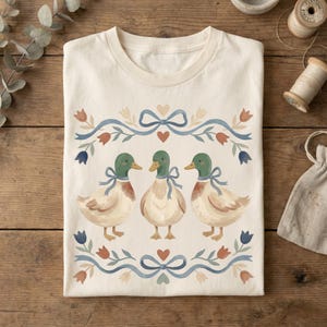 May include: Off-white t-shirt with a watercolour illustration of three ducks, each with green heads and blue ribbons. The design includes a floral border with blue ribbons, red and blue flowers, and hearts. The shirt is presented on a wooden surface.