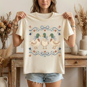 May include: A cream-coloured t-shirt displaying a design of three ducks with green heads and blue bows. The ducks are framed by a blue ribbon with floral and heart accents. The t-shirt is a casual, everyday wear item.