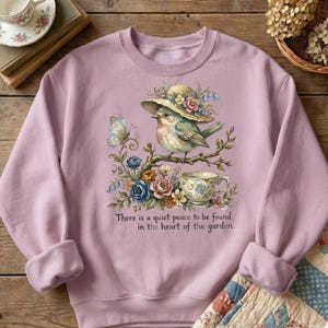 Pu&ograve; includere: Una felpa lilla con un'illustrazione ad acquerello di un uccello con un cappello, appollaiato su un ramo con fiori e una tazza da t&egrave;. Il testo recita: "C'&egrave; una quieta pace da trovare nel cuore del giardino."