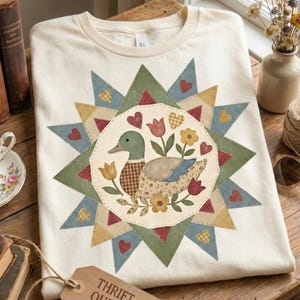 Peut inclure: T-shirt crème avec un motif de canard au centre. Le canard est entouré de fleurs et de cœurs dans un motif d'éclat d'étoile. L'éclat d'étoile a des accents verts, bleus et jaunes. Une étiquette avec le texte "THRIFT QUEEN" est visible.