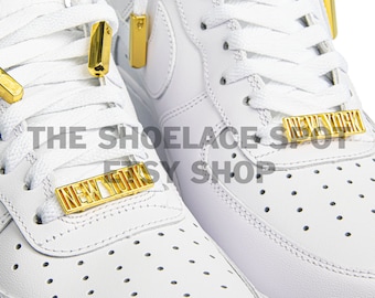 May include: White leather sneakers with gold metal lace tags that say "NEW YORK". The shoelaces are white and the sneakers have a white sole.