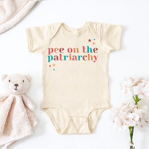 Könnte beinhalten: Ein cremefarbener Kurzarm-Babybody mit dem Text "pee on the patriarchy" in einer mehrfarbigen Schriftart. Das Design enthält kleine Sternakzente in den Farben Rot, Orange und Türkis. Das Kleidungsstück hat einen Rundhalsausschnitt und Druckknöpfe.