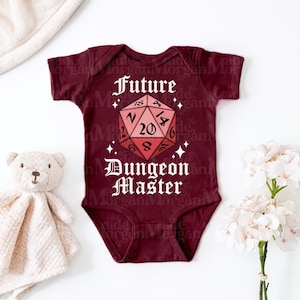 Könnte beinhalten: Ein kastanienbrauner kurzärmeliger Babybody mit einer Grafik eines rosafarbenen zwanzigseitigen Würfels, zentriert zwischen dem weißen Text Future und Dungeon Master. Das Design enthält kleine weiße Sternakzente um den Würfel auf der Vorderseite.