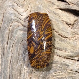 May include: A polished, oblong gemstone with a rich, earthy color palette. The stone features a dark brown base with intricate patterns of golden-yellow and black veining, creating a unique, natural design. The gemstone is likely used for jewelry or decorative purposes.