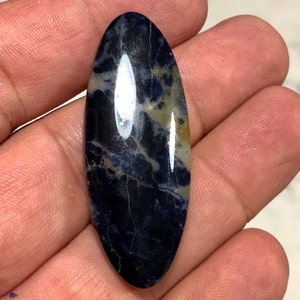 Puede incluir: Un cabuj&oacute;n liso de forma ovalada de sodalita azul y blanca con un patr&oacute;n de m&aacute;rmol azul oscuro y blanco.
