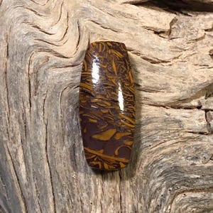 May include: A polished, rectangular gemstone with a unique pattern of brown and gold hues. The stone's surface reflects light, highlighting the intricate natural design. The gemstone is set against a textured wooden background.