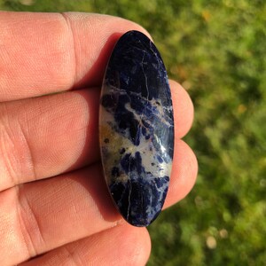 Puede incluir: Una piedra preciosa ovalada en cabuj&oacute;n con un patr&oacute;n de m&aacute;rmol en tonos azules y blancos. La piedra est&aacute; pulida y tiene una superficie lisa.