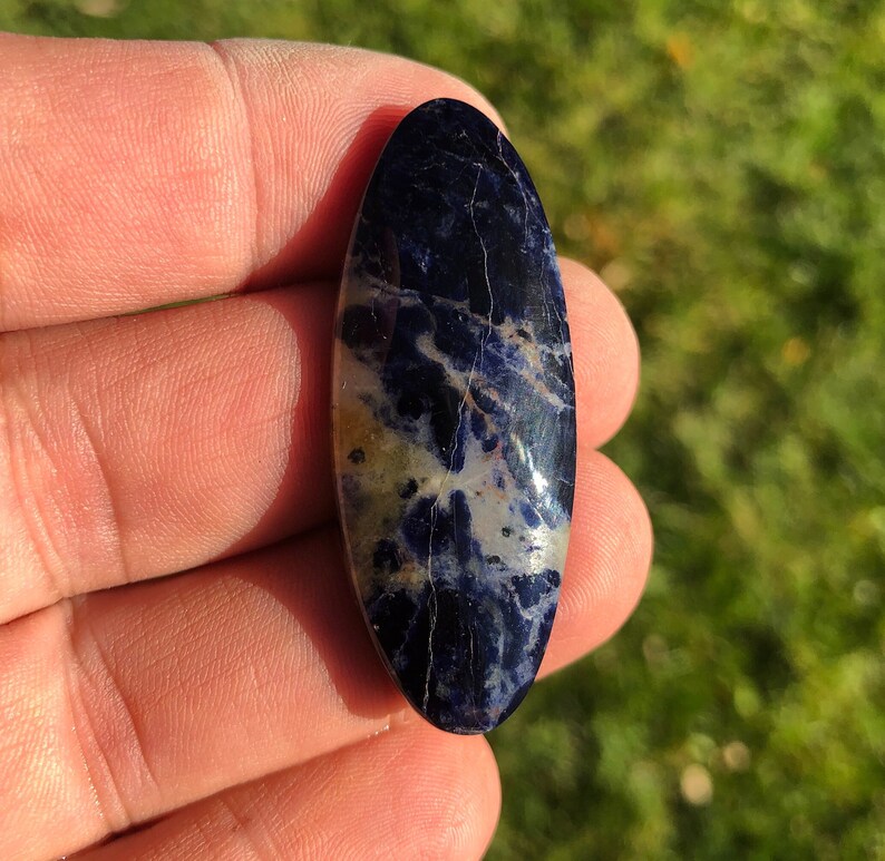Puede incluir: Una piedra preciosa ovalada en cabuj&oacute;n con un patr&oacute;n de m&aacute;rmol en tonos azules y blancos. La piedra est&aacute; pulida y tiene una superficie lisa.