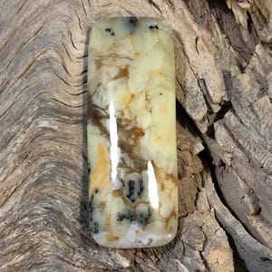 May include: A rectangular gemstone with a polished surface. The stone features a mix of colors, including creamy yellow, brown, and black, with a marbled pattern. The gemstone is set against a textured wooden background.