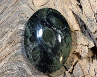 May include: Oval-shaped gemstone with a dark green and black pattern. The polished stone has a glossy finish and a unique, organic design. The gemstone is set against a textured, wooden surface.
