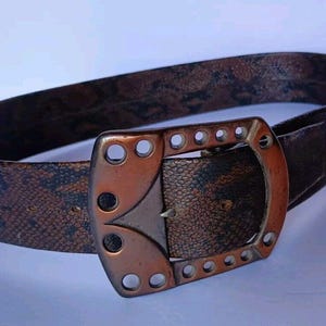 May include: A brown leather belt with a decorative, copper-toned buckle. The buckle has a unique, geometric design with multiple holes.