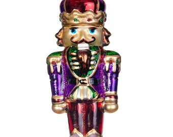 May include: A decorative Christmas ornament shaped like a nutcracker soldier. The ornament features a gold face with blue eyes and a black mustache. It wears a purple and red uniform with gold accents and a red crown. The ornament hangs from a gold string.