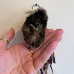 May include: A dark brown, textured fur keyring accessory featuring a small, rounded, plush-like form. Attached to the top is a silver-toned metal ring, and hanging from the bottom are several thin, multi-coloured fabric ribbons and strands.