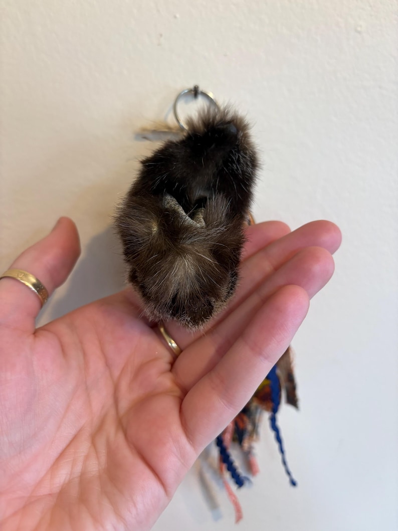 May include: A dark brown, textured fur keyring accessory featuring a small, rounded, plush-like form. Attached to the top is a silver-toned metal ring, and hanging from the bottom are several thin, multi-coloured fabric ribbons and strands.