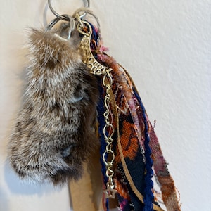 May include: A keyring featuring a brown and grey fur piece attached to a silver metal ring. A gold-toned hammered crescent moon charm hangs from a delicate chain with a small star, accompanied by several strips of multi-coloured patterned fabric.