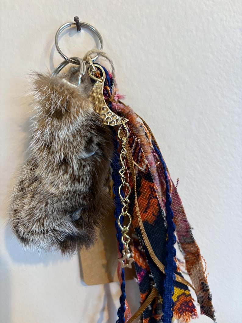 May include: A keyring featuring a brown and grey fur piece attached to a silver metal ring. A gold-toned hammered crescent moon charm hangs from a delicate chain with a small star, accompanied by several strips of multi-coloured patterned fabric.