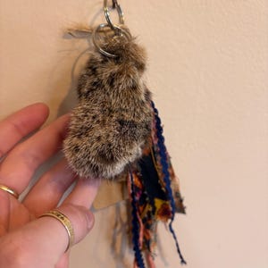 May include: A keyring featuring a fluffy, brown and black mottled fur charm attached to a silver metal ring. The charm is accented with a cluster of hanging fabric strips in navy blue, orange, and yellow floral patterns with frayed edges.